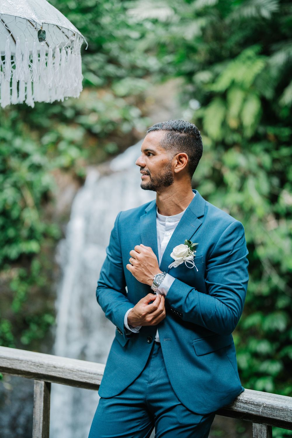 Groom in blue suit adjusts his cuffs while standing outdoors near greenery.
