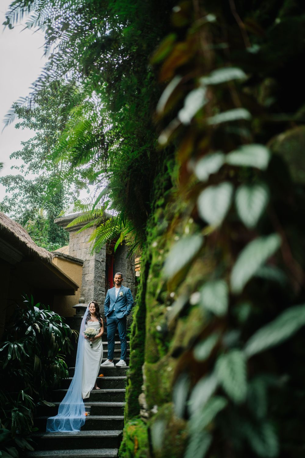 A couple in wedding attire walking down stone steps surrounded by lush foliage.