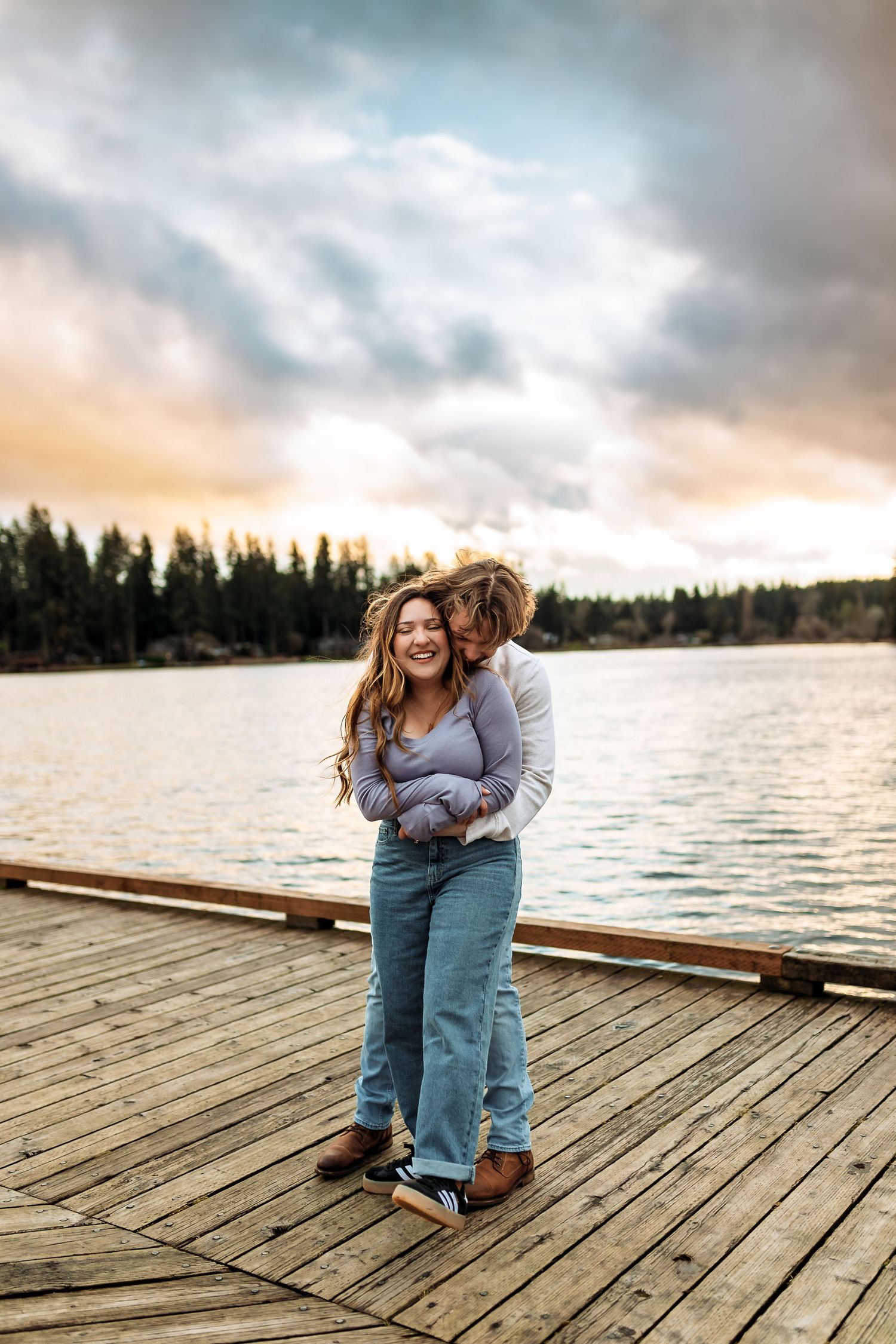 Couple embracing on wooden dock during sunset with evergreen forest and lake in background.