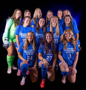 Women's soccer team poses in blue uniforms and one green goalkeeper uniform against dark background with dramatic lighting.