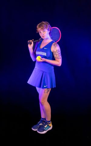 Tennis player poses with racquet in blue Nike dress against dramatic dark background.