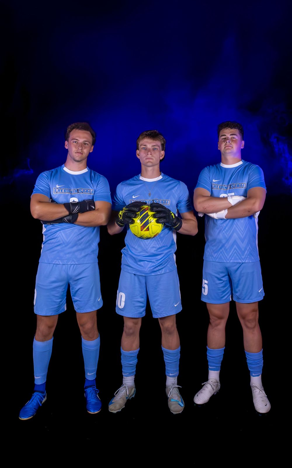Soccer team members in light blue uniforms pose together against dark background with dramatic lighting.