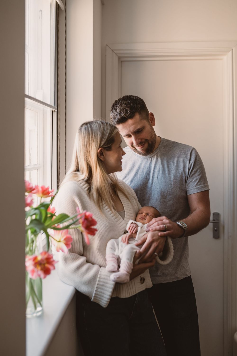 A couple stands by a window with their newborn baby in a bright, minimalist interior with pink flowers in the foreground.
