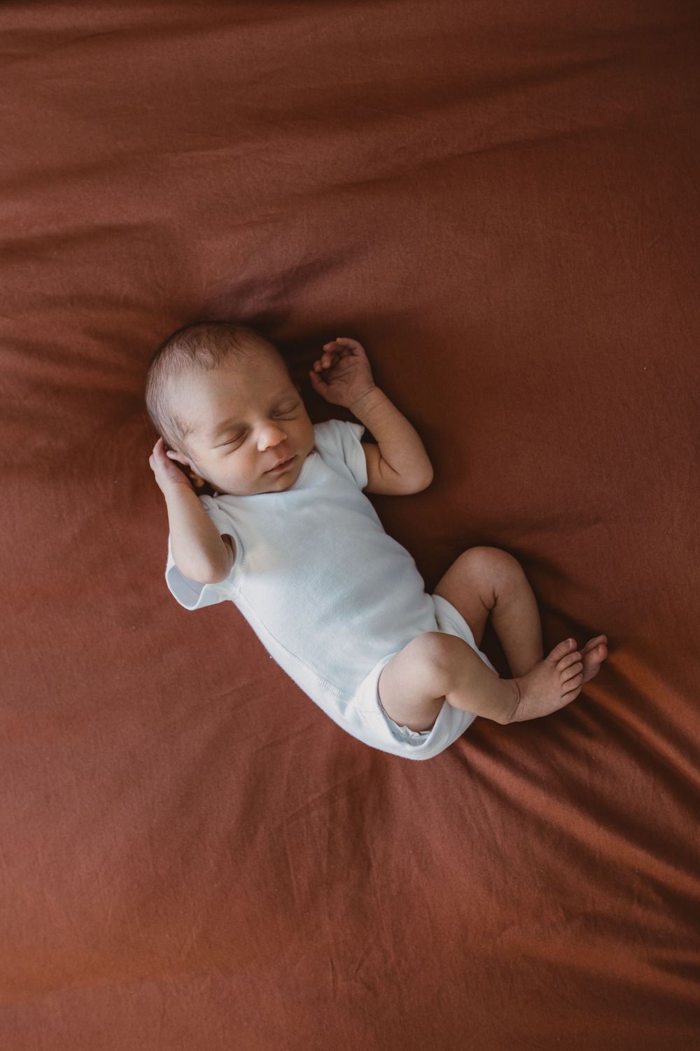 A sleeping newborn baby in a white vest lies on rust coloured bedding.
