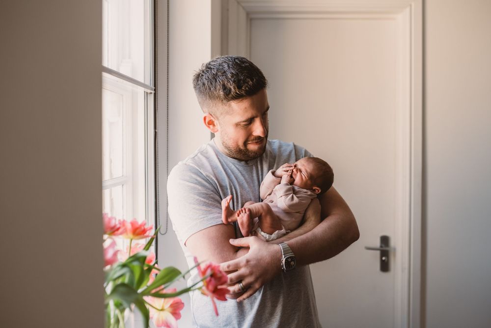 A father in a gray t-shirt tenderly holds and gazes at their newborn baby near a window with pink tulips.