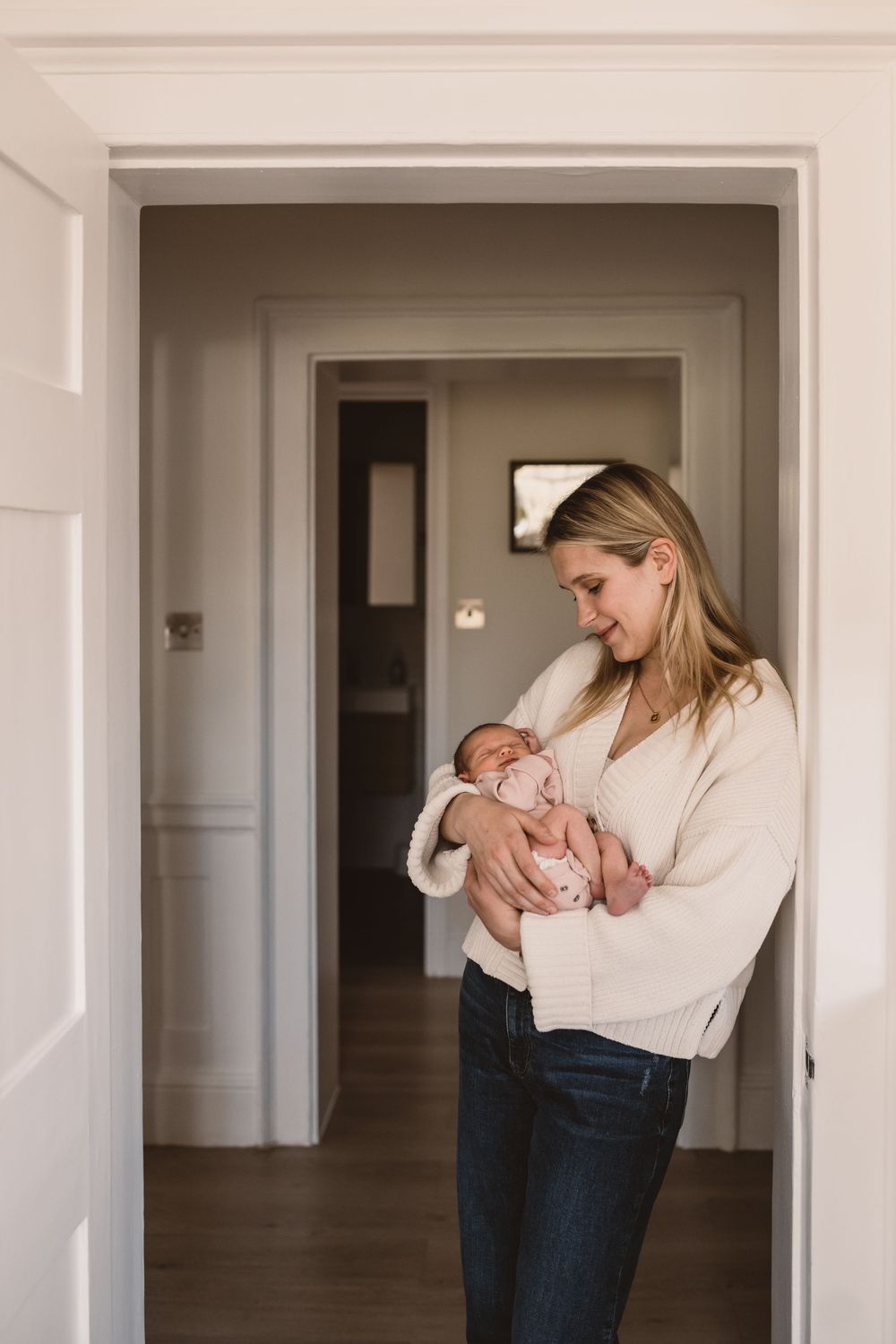 A mother in a cream sweater holds a newborn baby while standing in a doorway of a modern home.