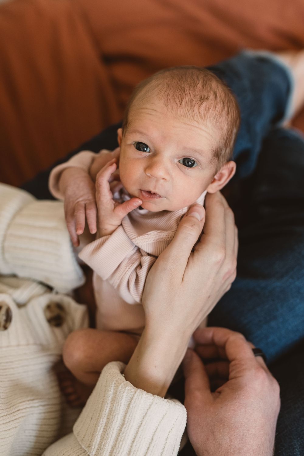 A newborn baby wearing a light pink outfit is being gently cradled by hands in cream colored sleeves.