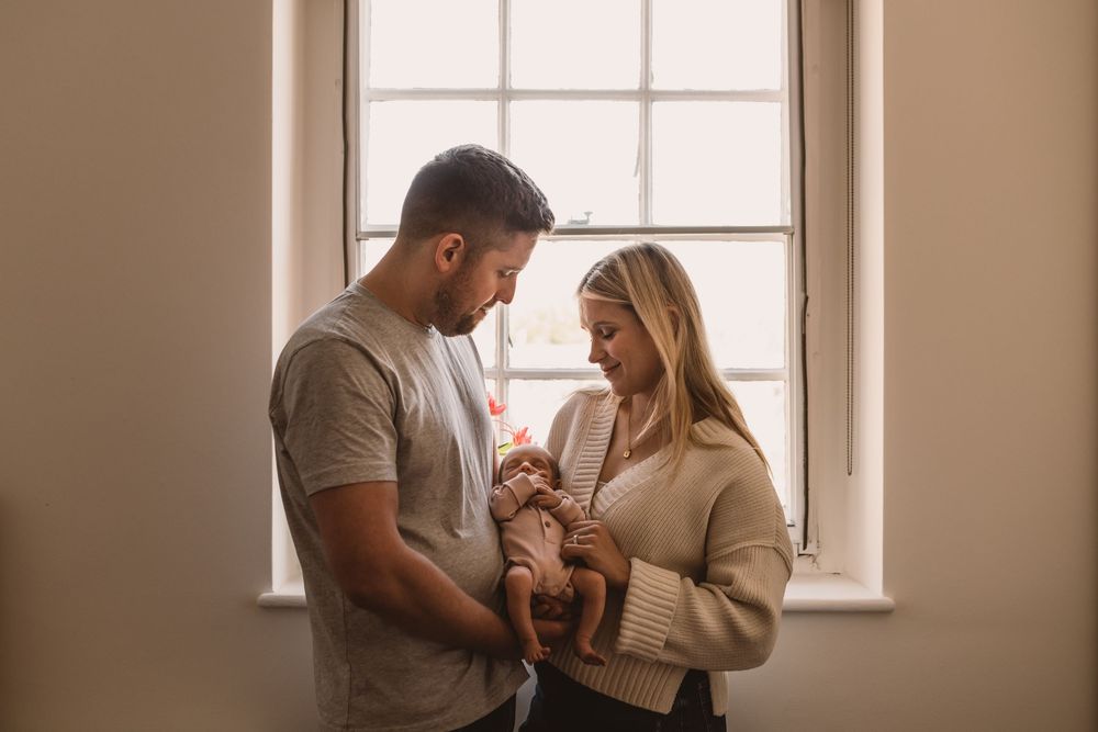 Couple in warm lighting by window shares intimate moment while holding their newborn baby.