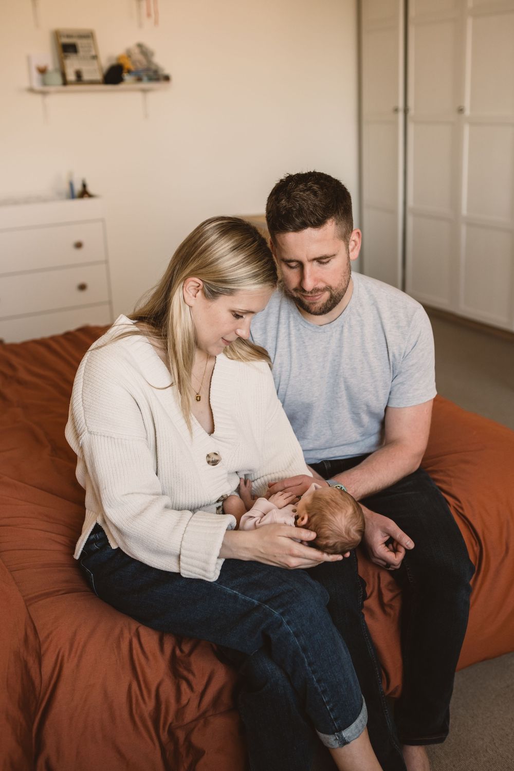 Couple sitting together on brown bedding holding their newborn baby in minimalist white room.