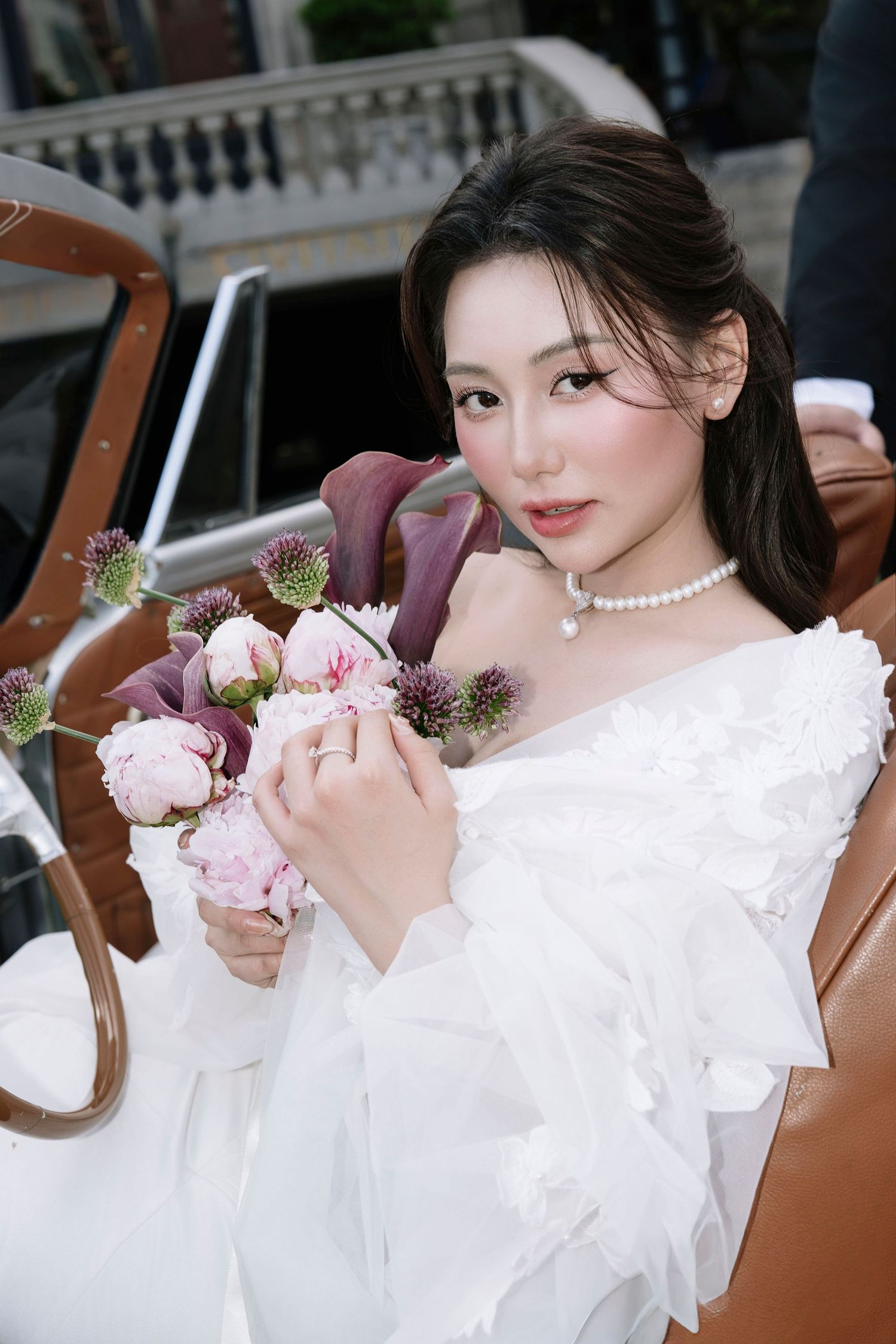 Beautiful Asian bride in white ruffled gown holds pink and purple floral bouquet beside vintage car.