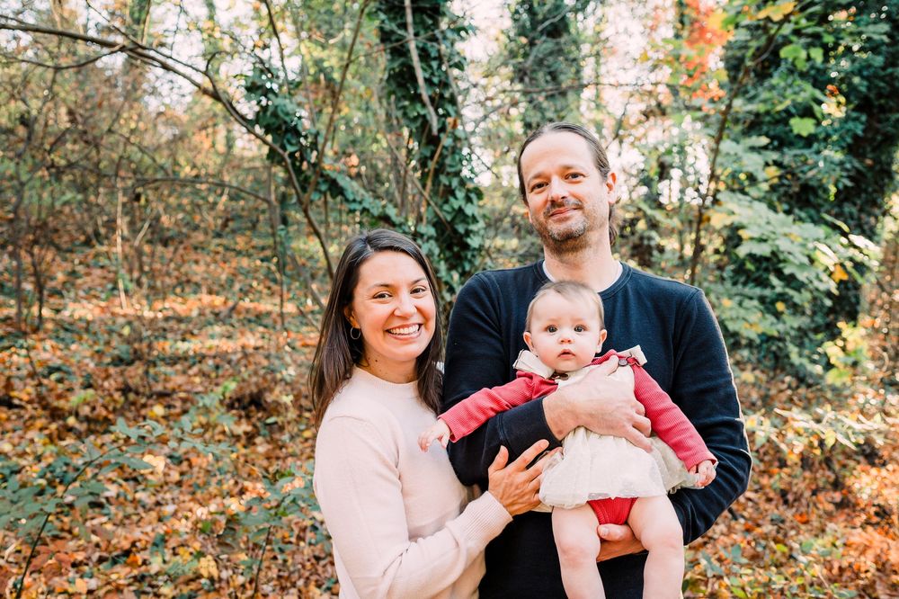 Family in casual attire posing for autumn portraits among colorful fall foliage in an outdoor park setting.