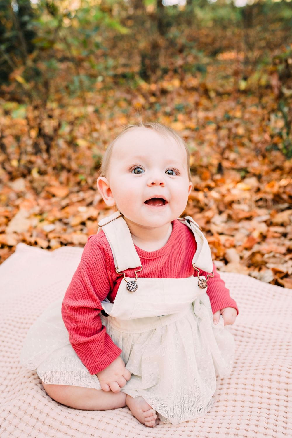 A sweet baby sits on a blanket surrounded by fallen autumn leaves in a warm outdoor setting.