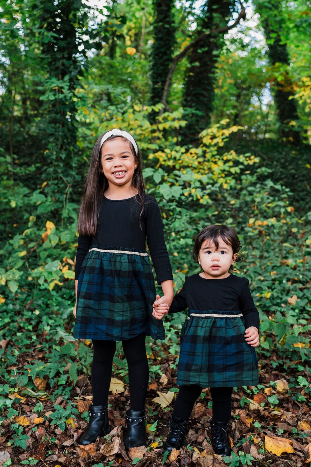 Two children in matching navy plaid dresses play among autumn leaves and greenery in a wooded setting.