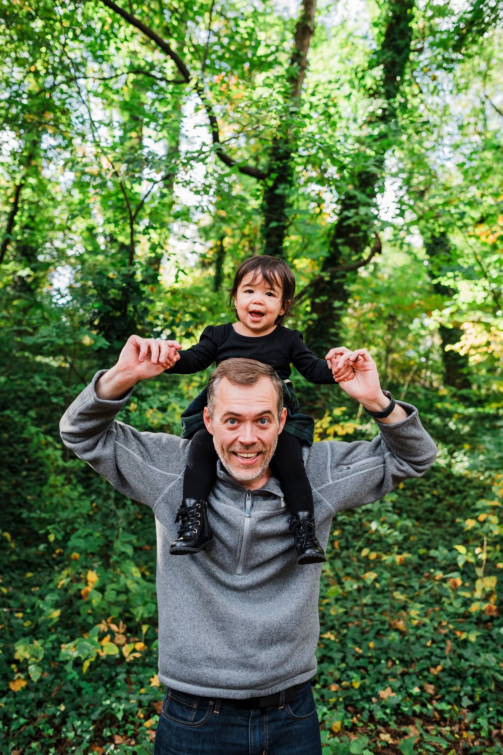 A happy young child rides on someone's shoulders in a lush green forested area during autumn.