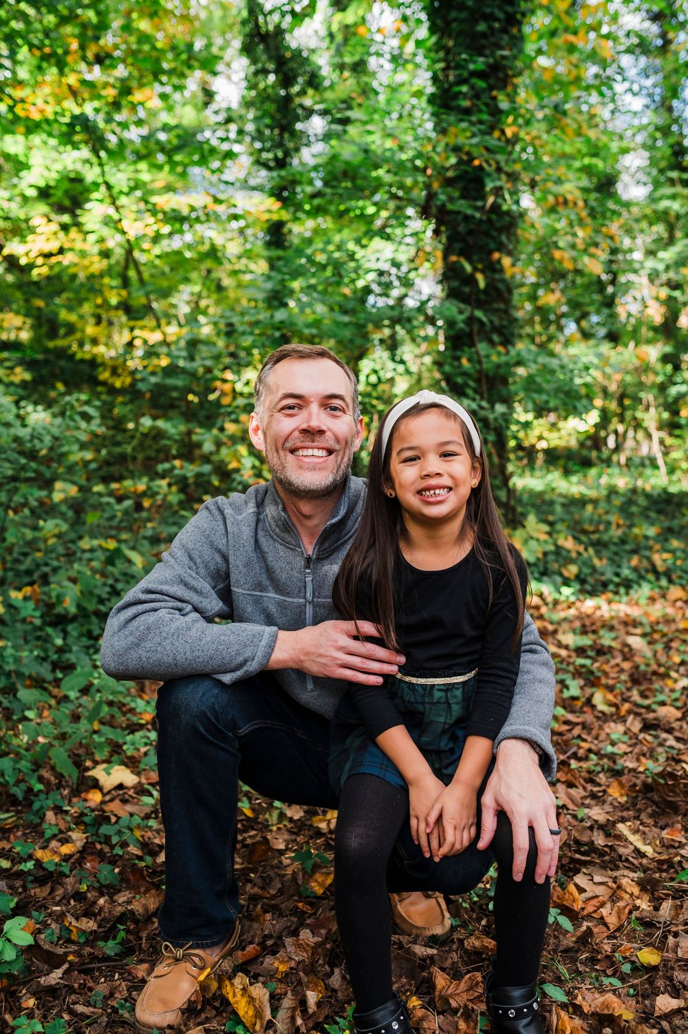 A happy parent and child sit together on fallen autumn leaves in a forest setting during a fall photo session.