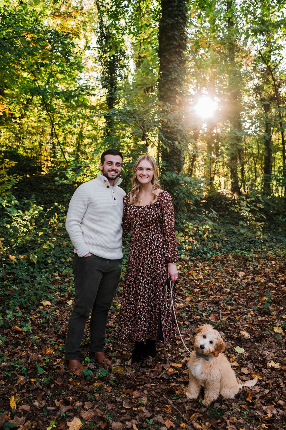 Golden sunlight streams through autumn forest trees as a couple poses with their fluffy goldendoodle dog on leaf-covered ground.