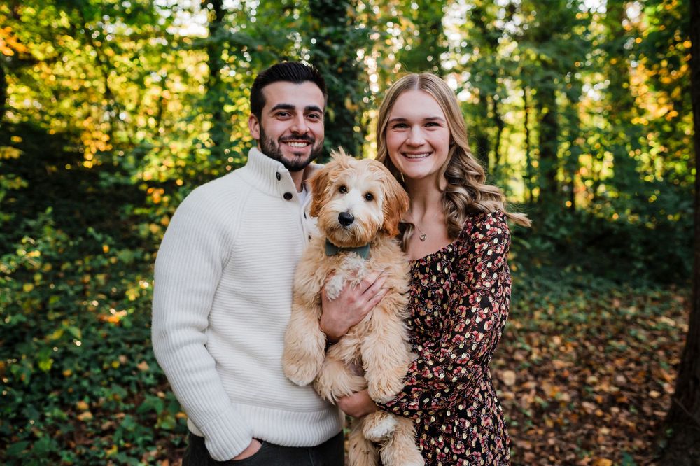 A couple poses outdoors during an autumn engagement photo session with their golden doodle puppy in a wooded area.
