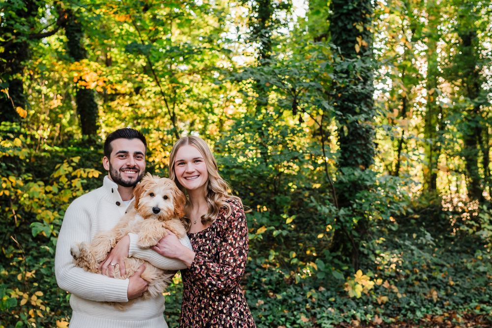A young couple poses with their red-haired dog in a sunlit autumn forest setting.