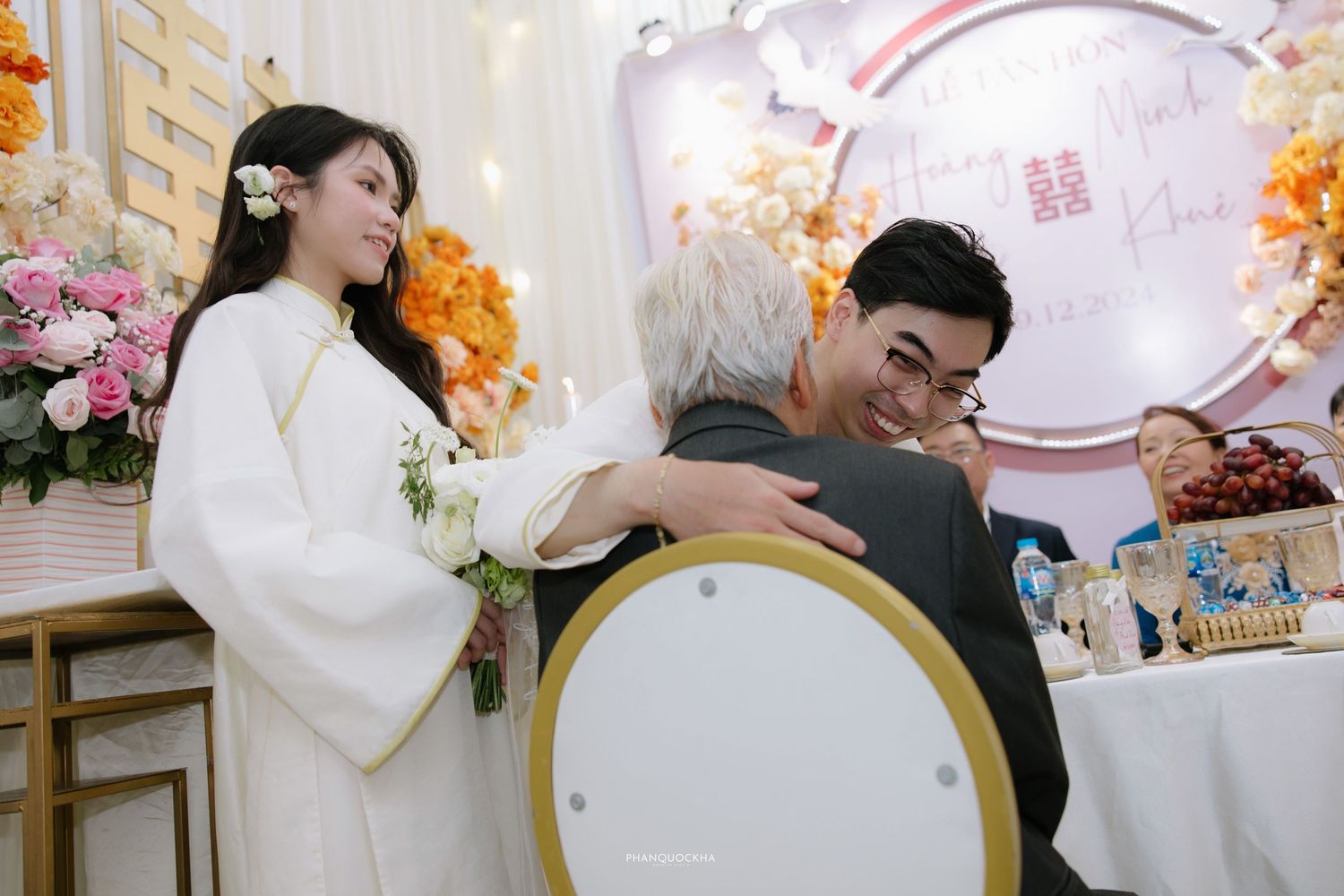 Groom in white ao dai embraces elderly guest while bride stands nearby at Vietnamese wedding.