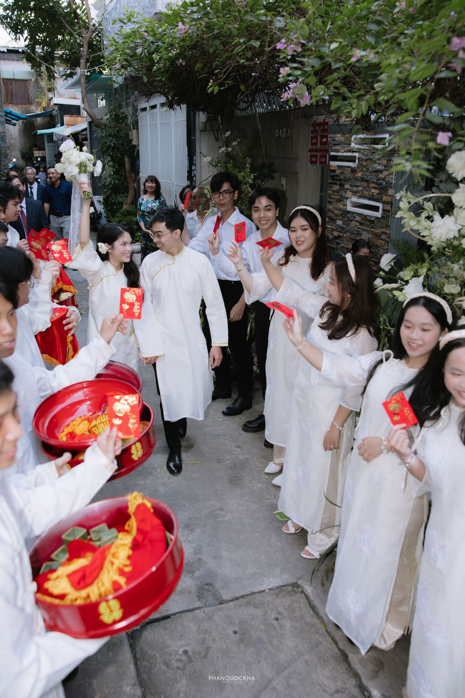 Vietnamese wedding procession with guests in white ao dai holding red envelopes and ceremonial trays outdoors.