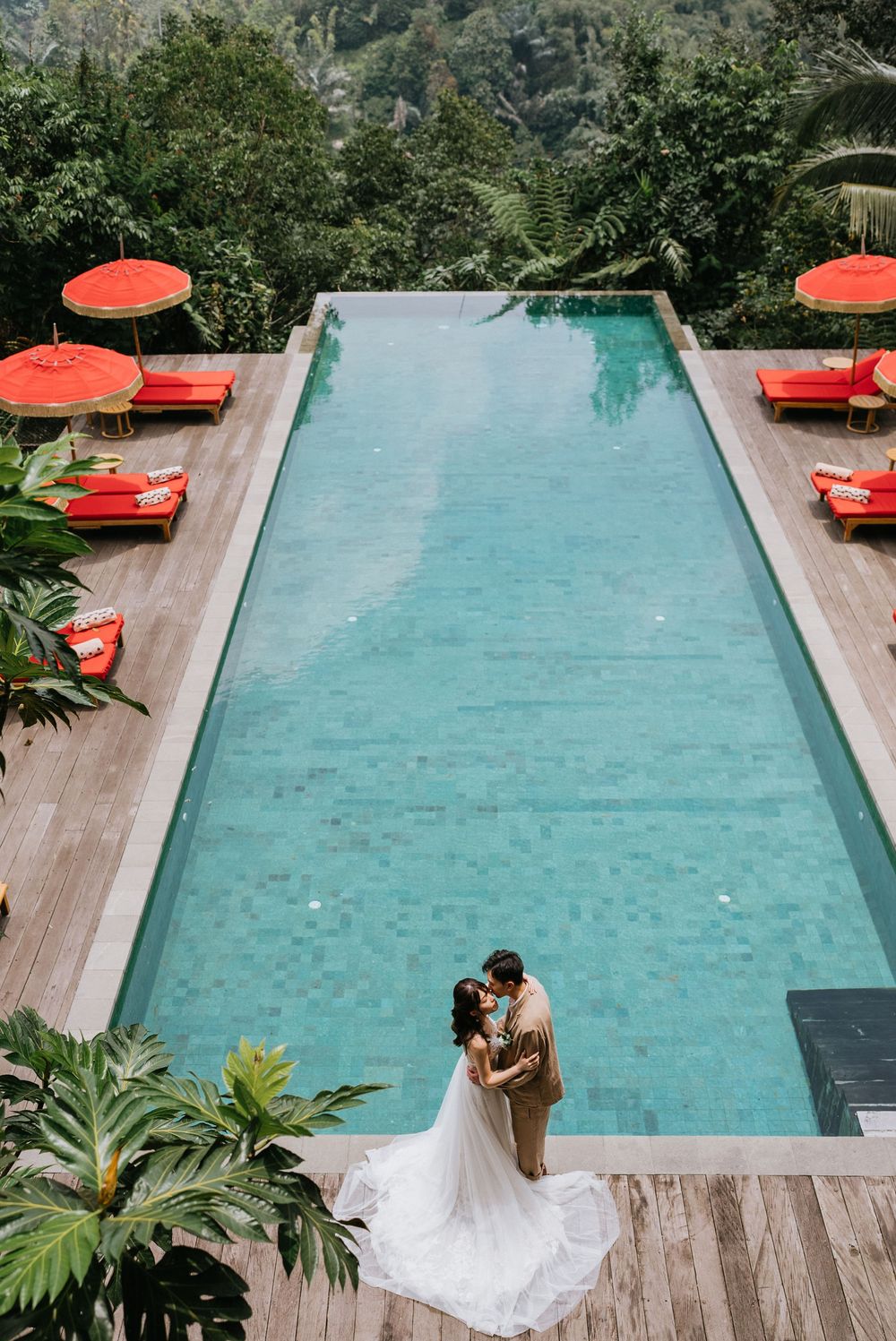 A tropical poolside setting with red umbrellas and wooden deck surrounded by lush greenery.
