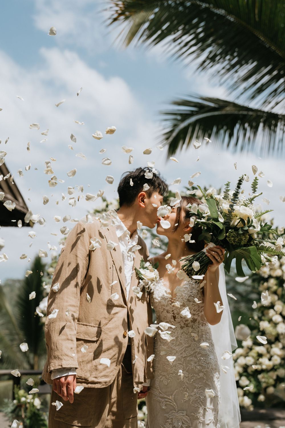 Wedding couple celebrating outdoors under palm trees with flower petals being tossed in the air.