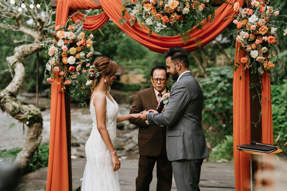 Wedding ceremony under a vibrant orange draped arch decorated with white and peach flowers in an outdoor setting.