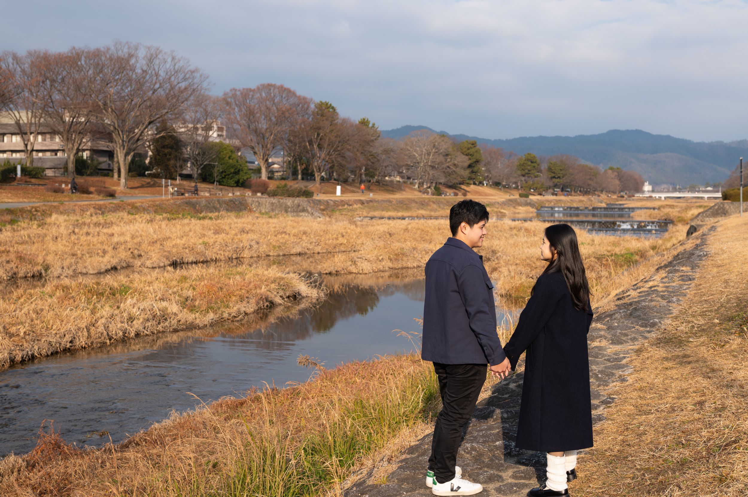 Couple holding hands on a dirt path beside a narrow stream with dry golden grass in winter.