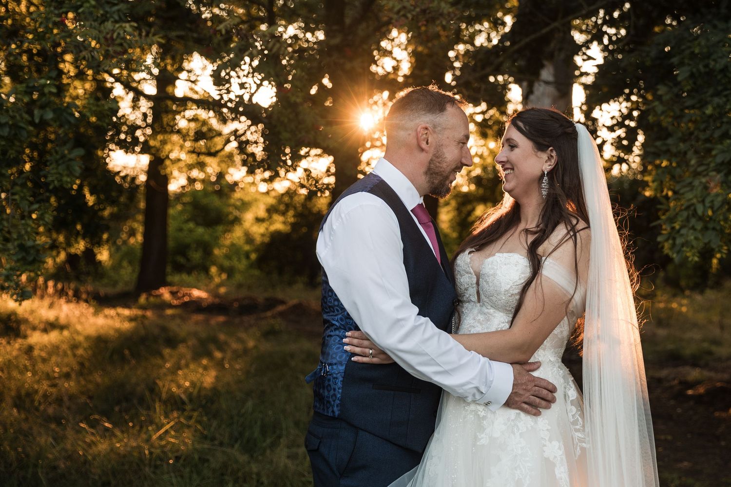 Bride and groom embracing in a romantic outdoor setting with soft sunlit trees and foliage at Kelham House