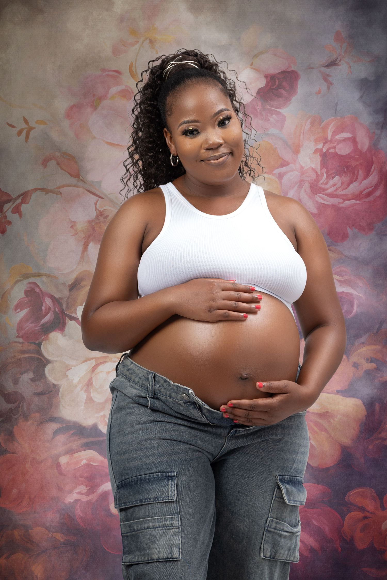 An expectant mother in white tank top and cargo pants poses against a floral backdrop.