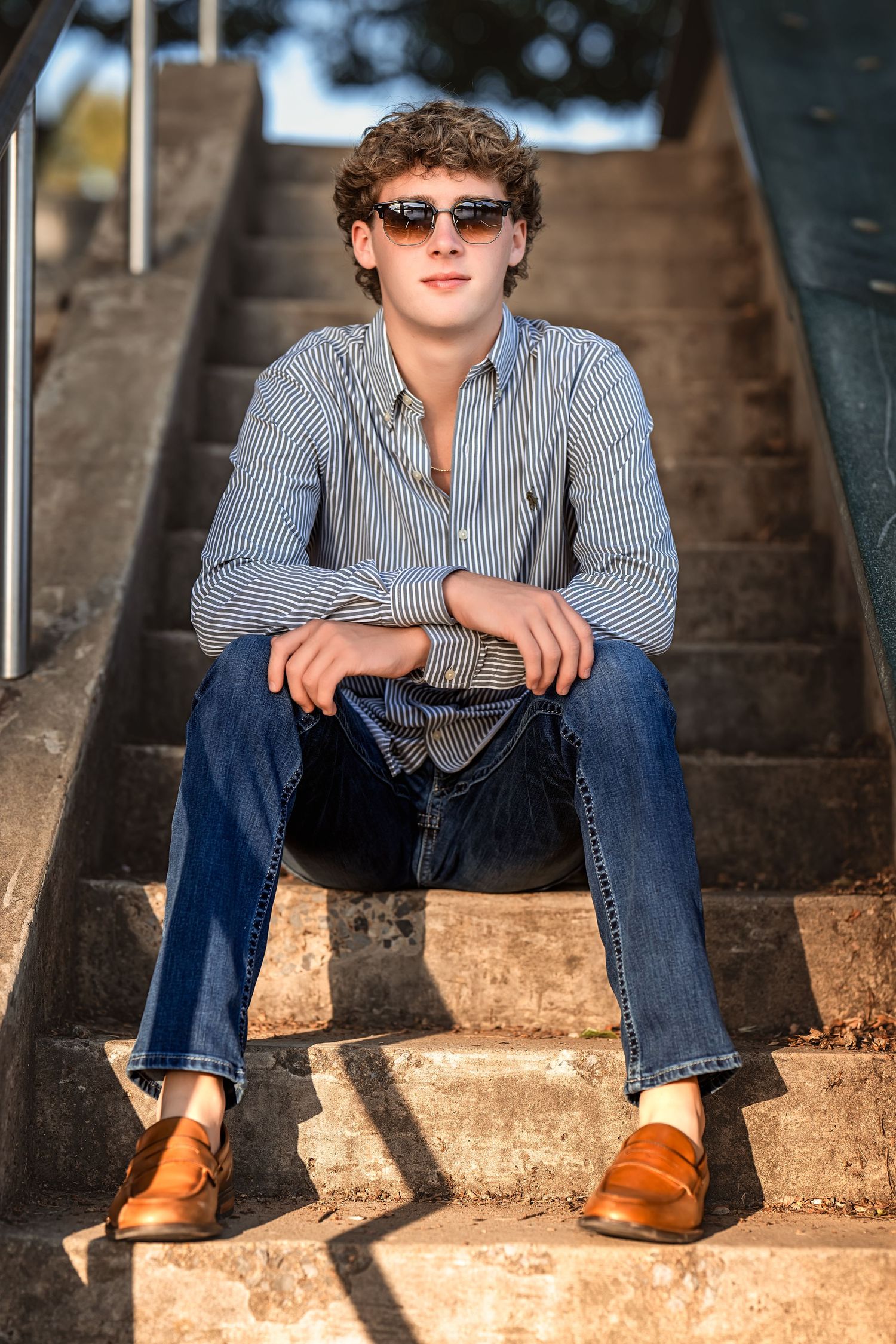 Senior boy in sunglasses sitting on concrete steps wearing a striped shirt, blue jeans, and brown suede shoes in outdoor lighting.