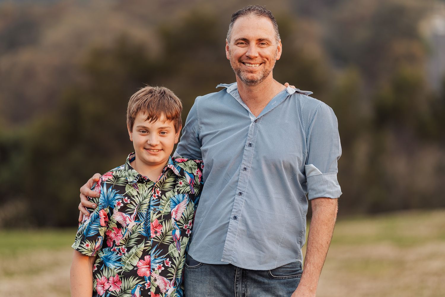 A parent and child stand together outdoors smiling at the camera, with the child wearing a colorful floral shirt.