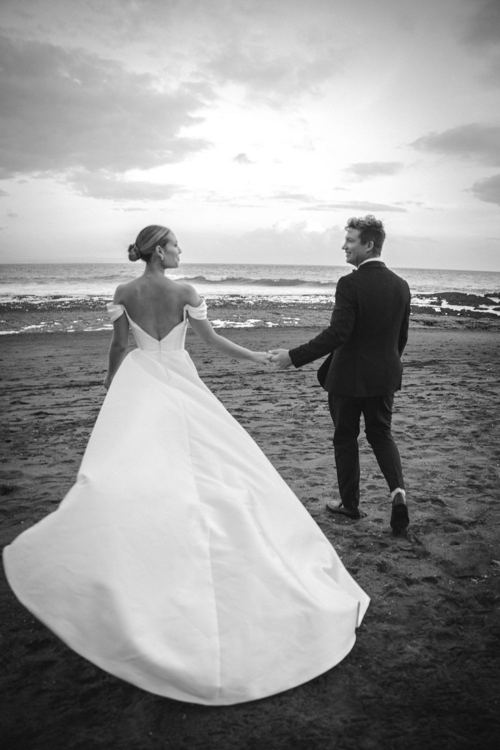A romantic black and white photo of a couple on a beach with the bride's white dress flowing in the wind at sunset.