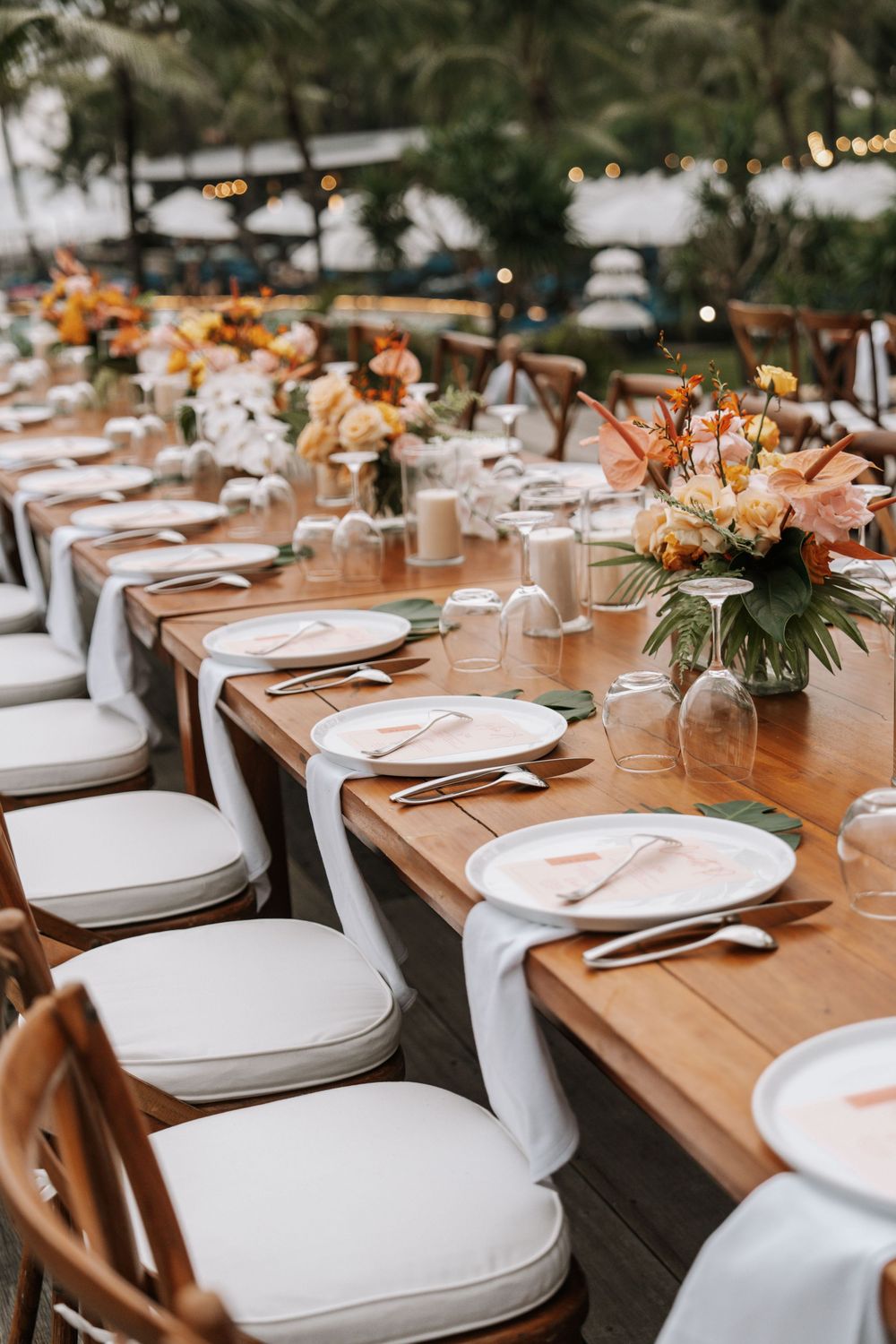 Long wooden tables set for a rustic outdoor wedding reception with white plates and chairs at sunset.
