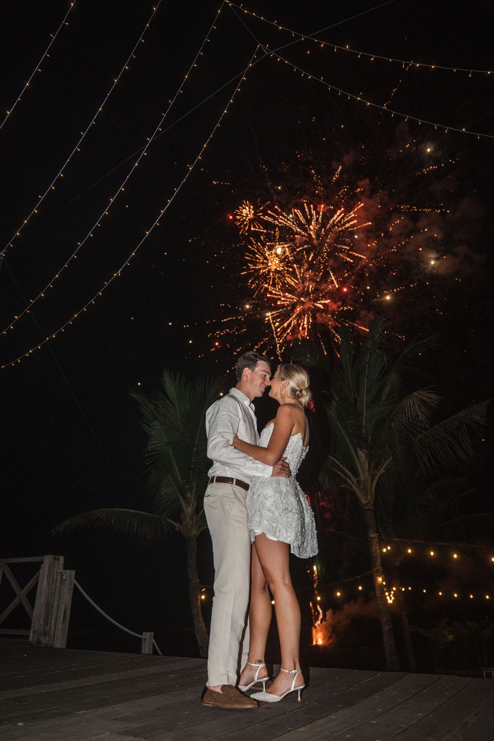 A couple shares a romantic moment on a deck under string lights and fireworks at night, with palm trees in the background.