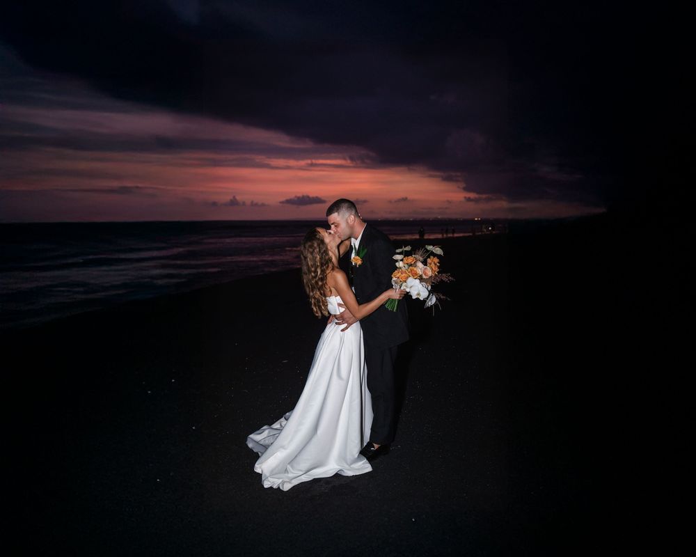 Two people share a romantic kiss on a beach at sunset with dramatic purple clouds overhead while wearing wedding attire.