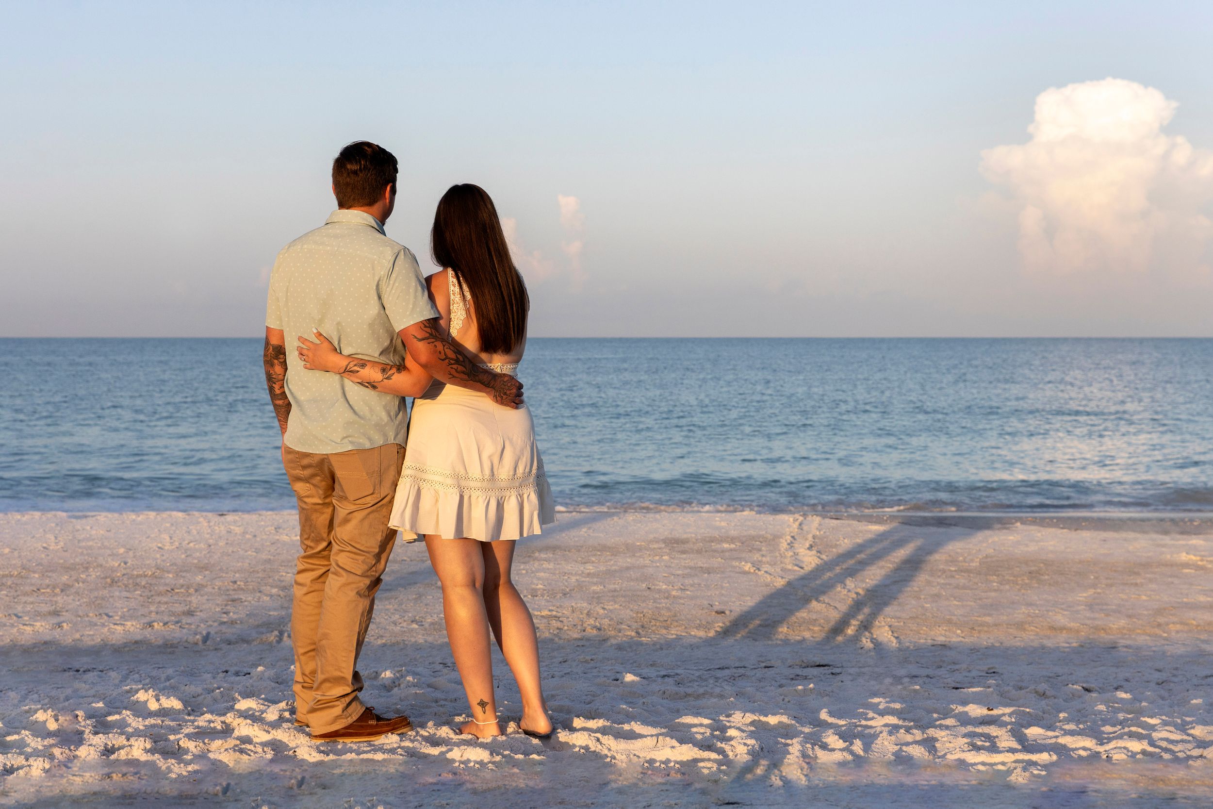 Couple standing together on a sandy beach at sunset, gazing at the calm ocean horizon in a romantic seaside moment.