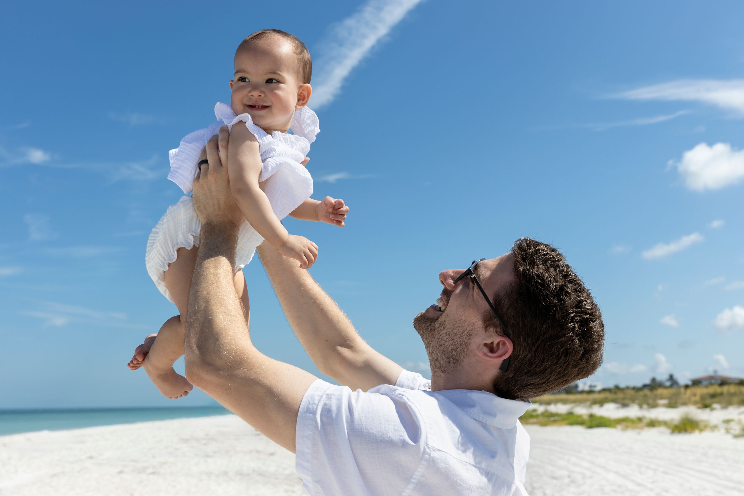 Father joyfully lifting smiling baby girl in white dress on sunny tropical beach with blue sky.