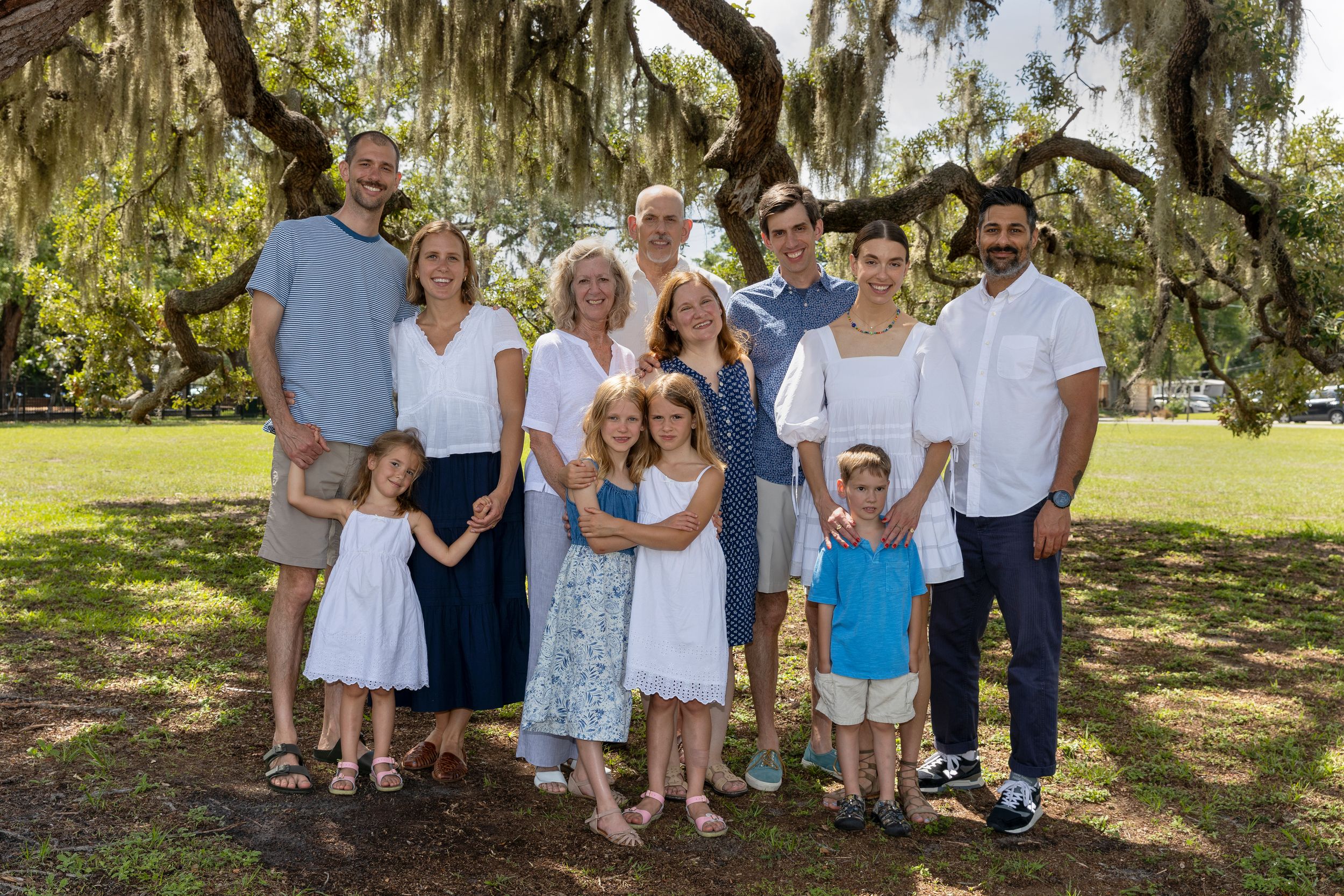 Large family group portrait outdoors under sprawling oak trees, adults and children smiling in coordinated blue and white outfits.