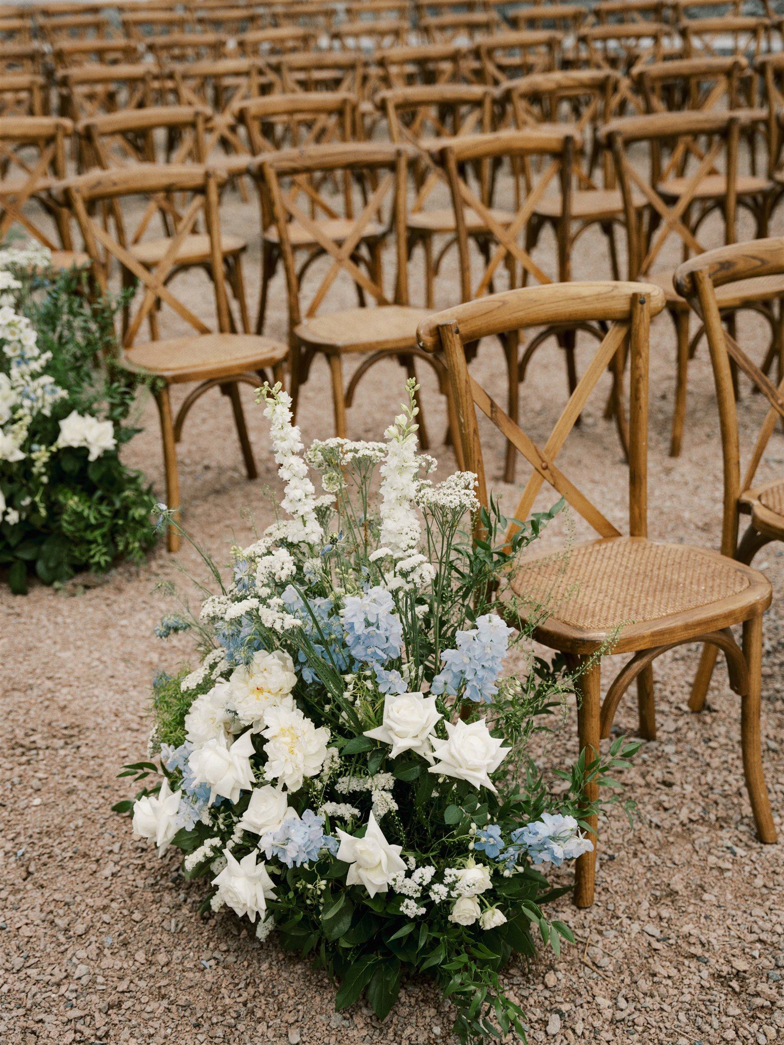 Rustic wooden crossback chairs arranged in rows with delicate white and blue floral arrangements on gravel ground.