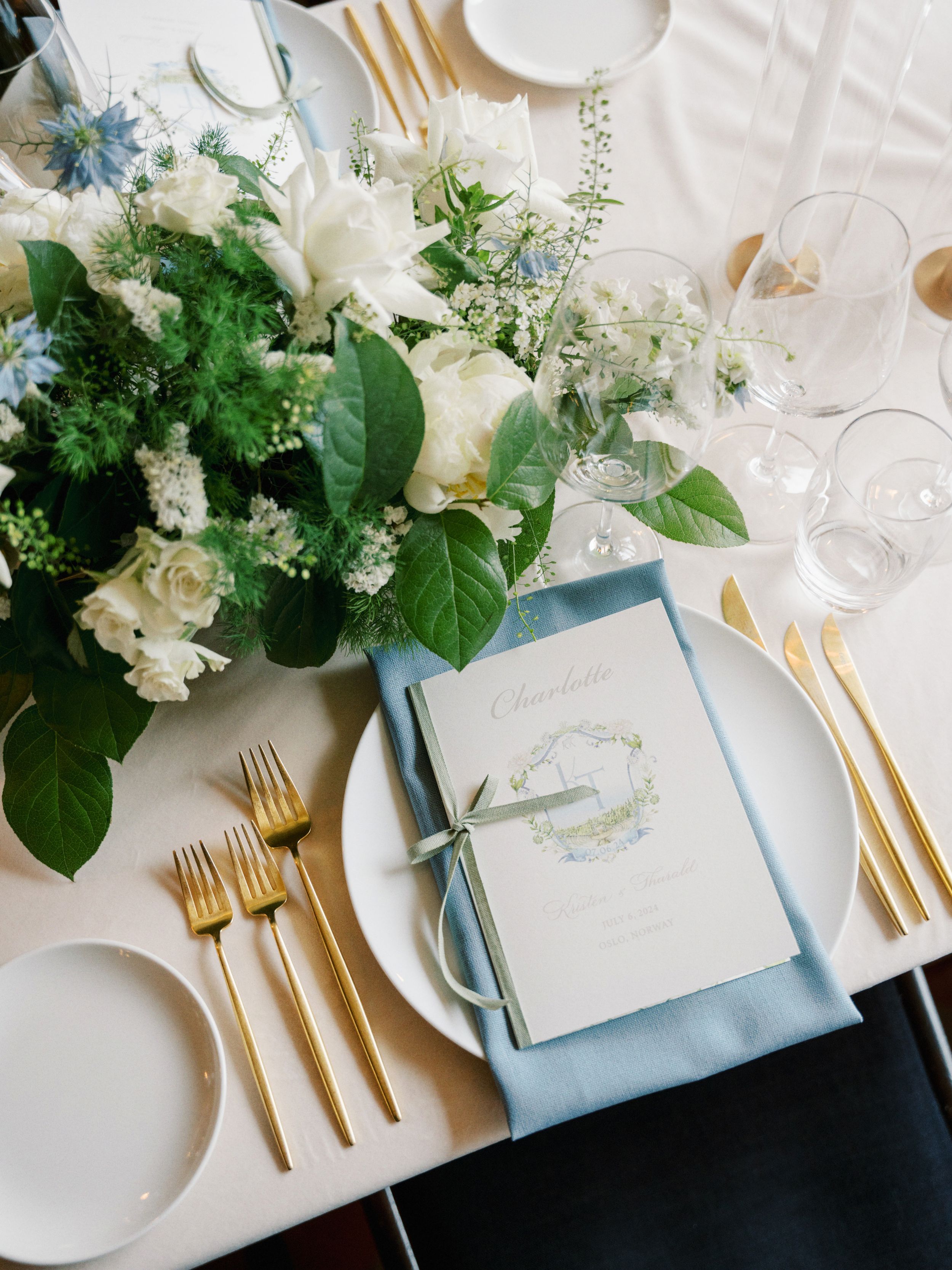 Elegant wedding table settings with light blue napkins, gold cutlery, and white floral centerpieces arranged on a white tablecloth.