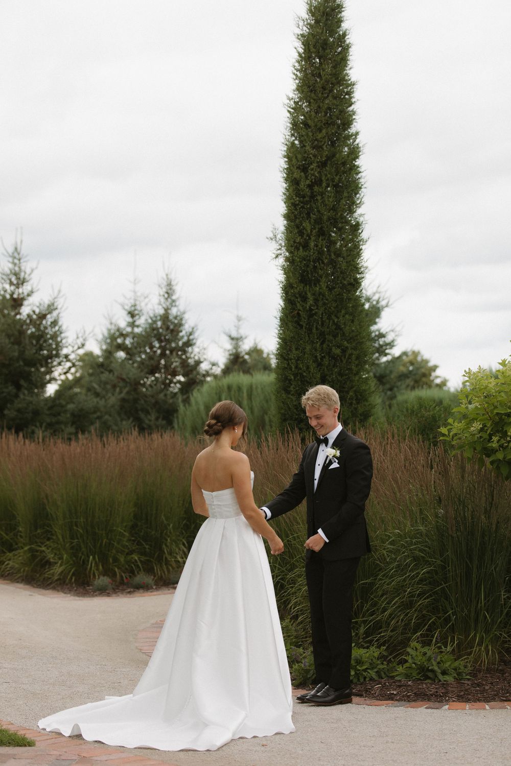 Couple in formal wedding attire standing on a garden path with tall cypress trees in background.