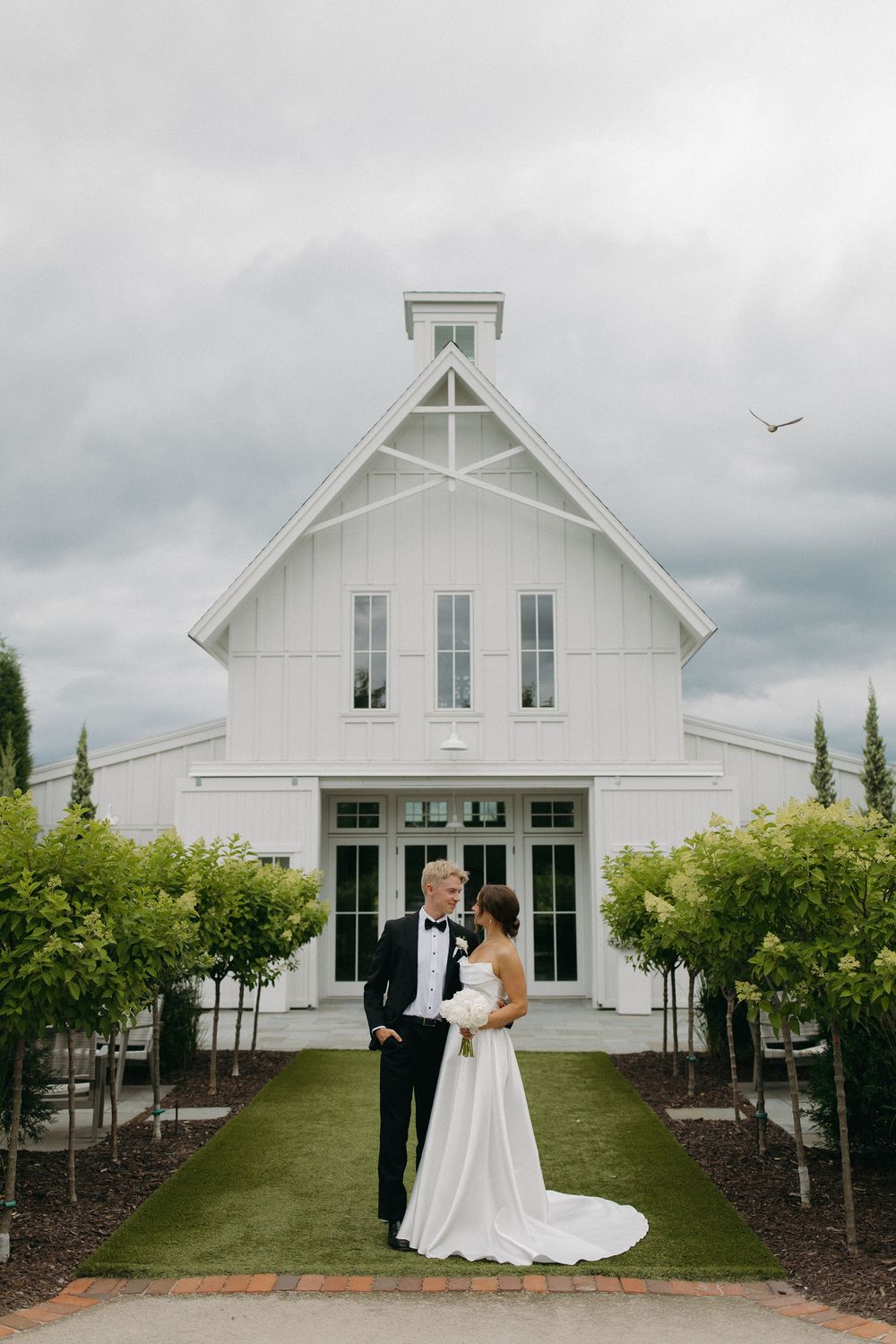 Wedding couple poses in front of a modern white farmhouse building with landscaped grounds.