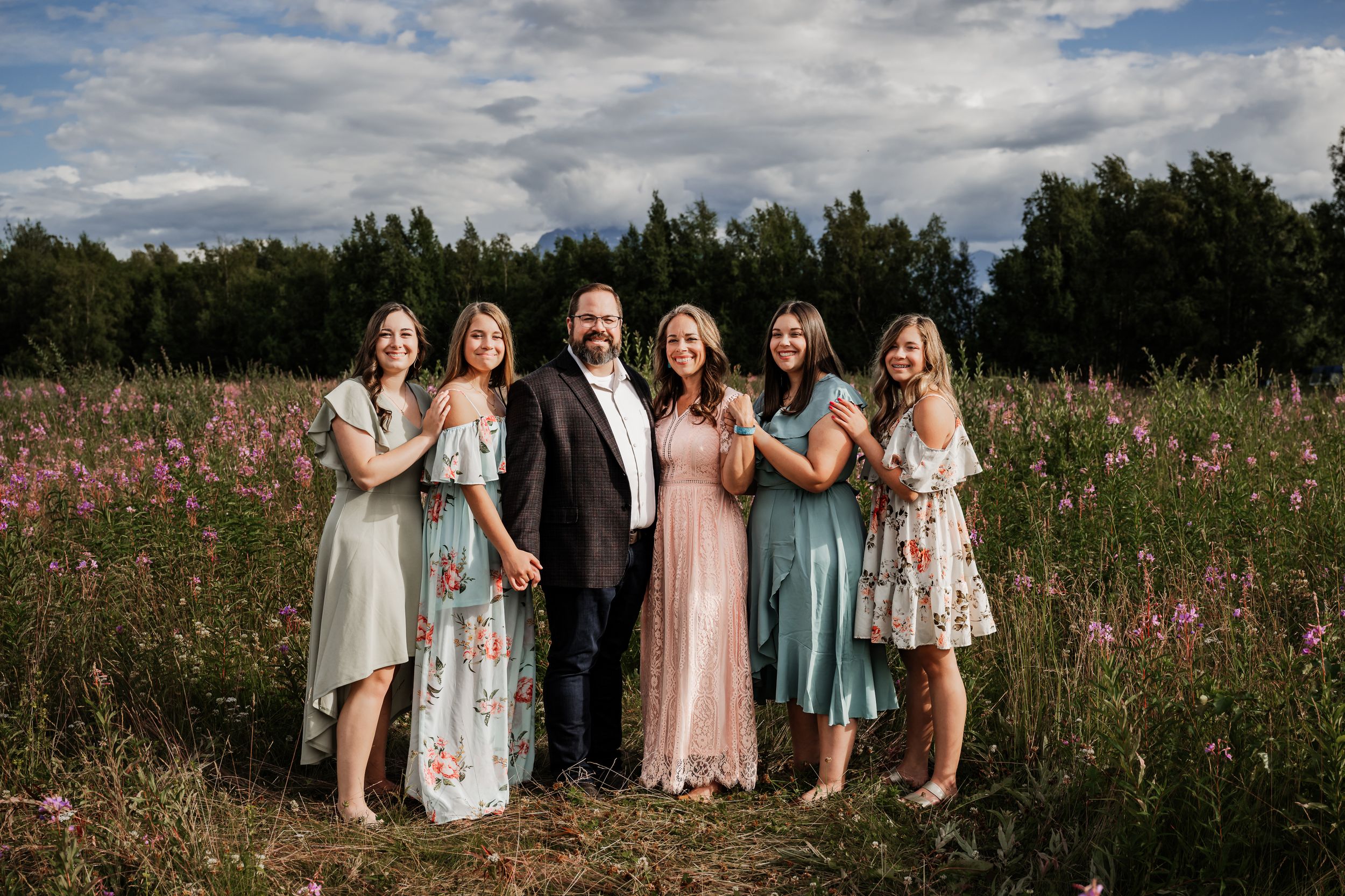 Family of 6 standing in a wildflower meadow with pink blooms under a cloudy summer sky.