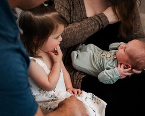 Lifestyle in home born family photos. Older sister sits in her fathers lap as she stares at her new baby brother.