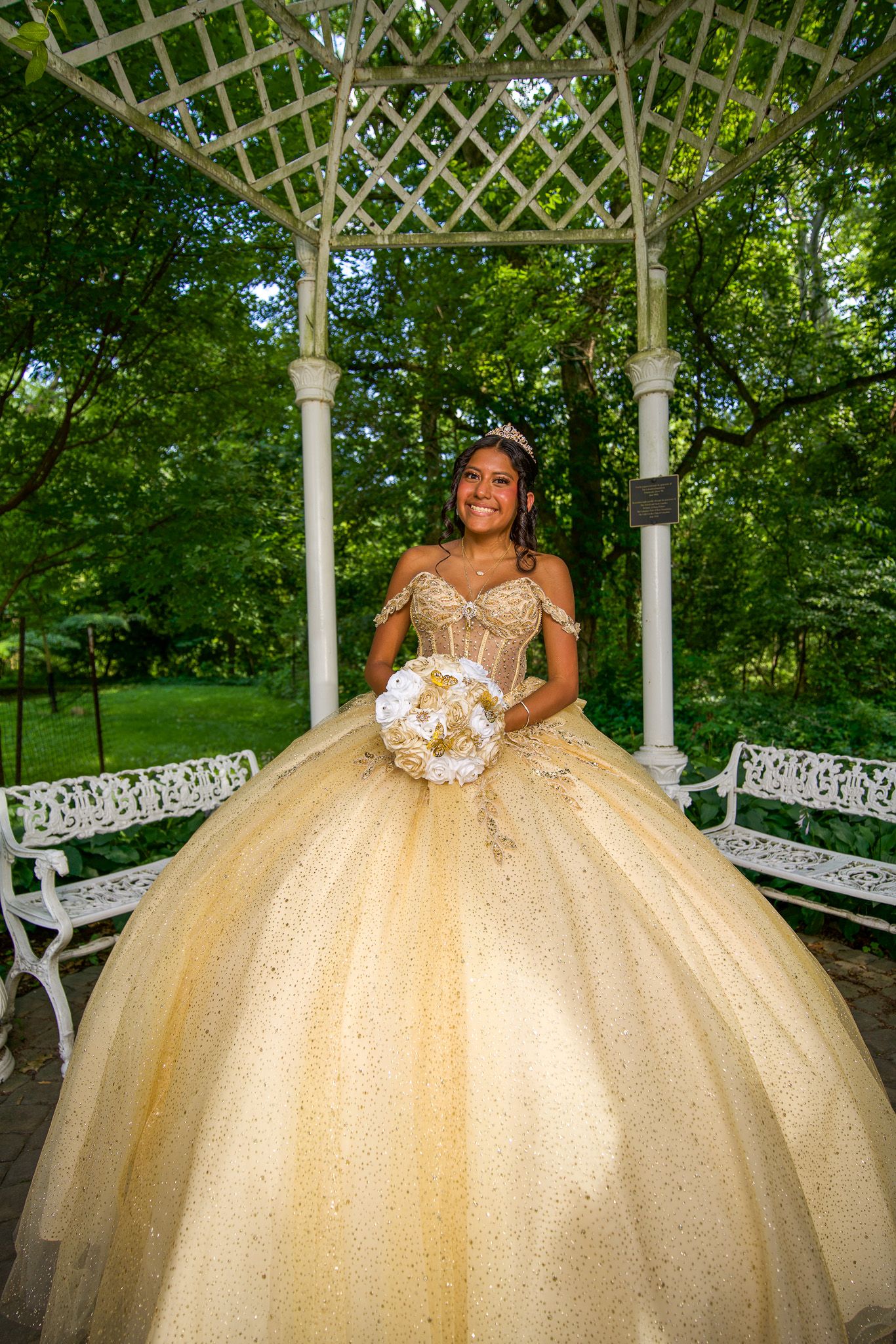 QUINCEAÑERA Stunning gold formal dress photographed in a white garden gazebo with intricate lattice details.