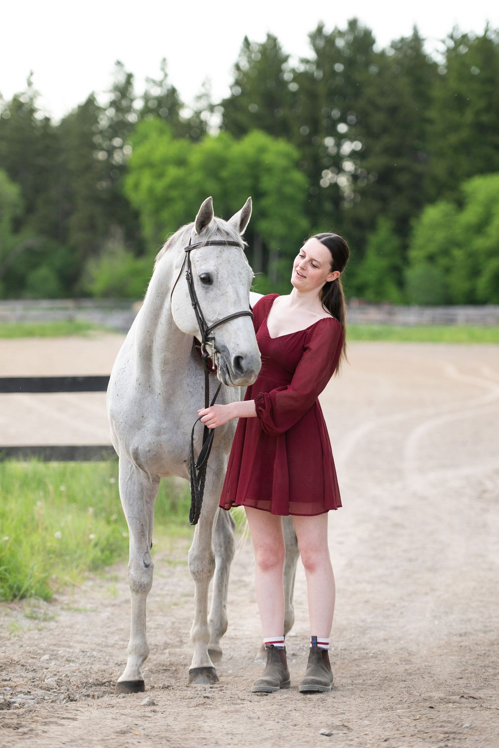 A woman in a burgundy dress lovingly interacts with a beautiful white horse on a dirt path surrounded by greenery.