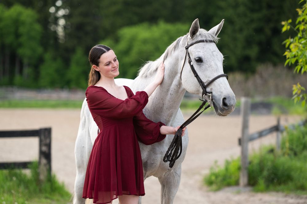 A woman in a burgundy dress poses with a beautiful white horse in an outdoor setting during a photo shoot.