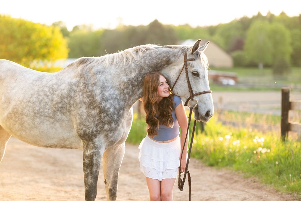 A dappled gray horse and a woman in white stand in a sunlit meadow with yellow wildflowers.