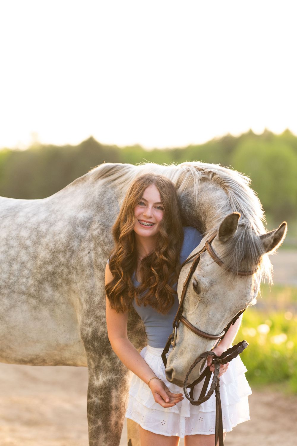 A grey dappled horse and person in white dress share a tender moment at golden hour in a meadow.